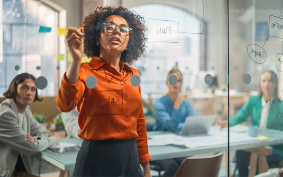 A Professional Black Business Woman Explains a Strategic Plan to a Diverse Team of People Using a Glass Board and a Marker.